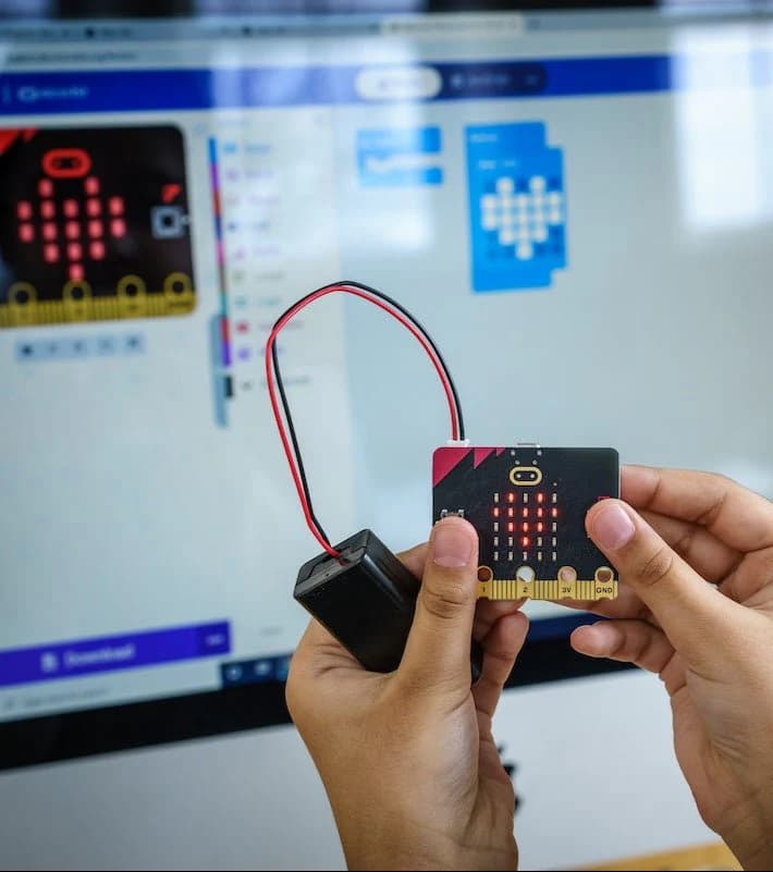 Hands holding a micro:bit with battery holder in front of a large monitor